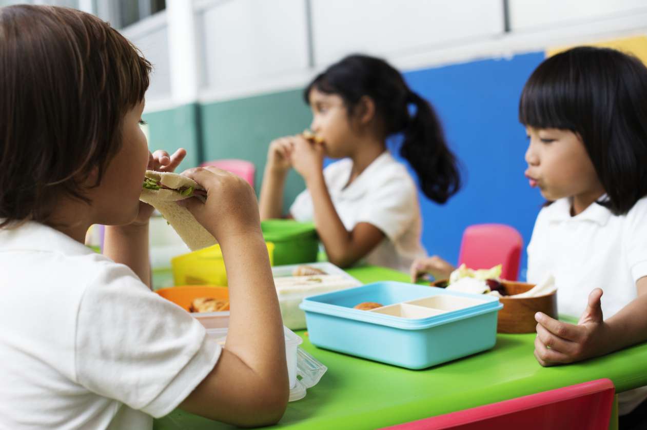 Three children sit at a lunch table at school and eat their lunches out of colourful containers.