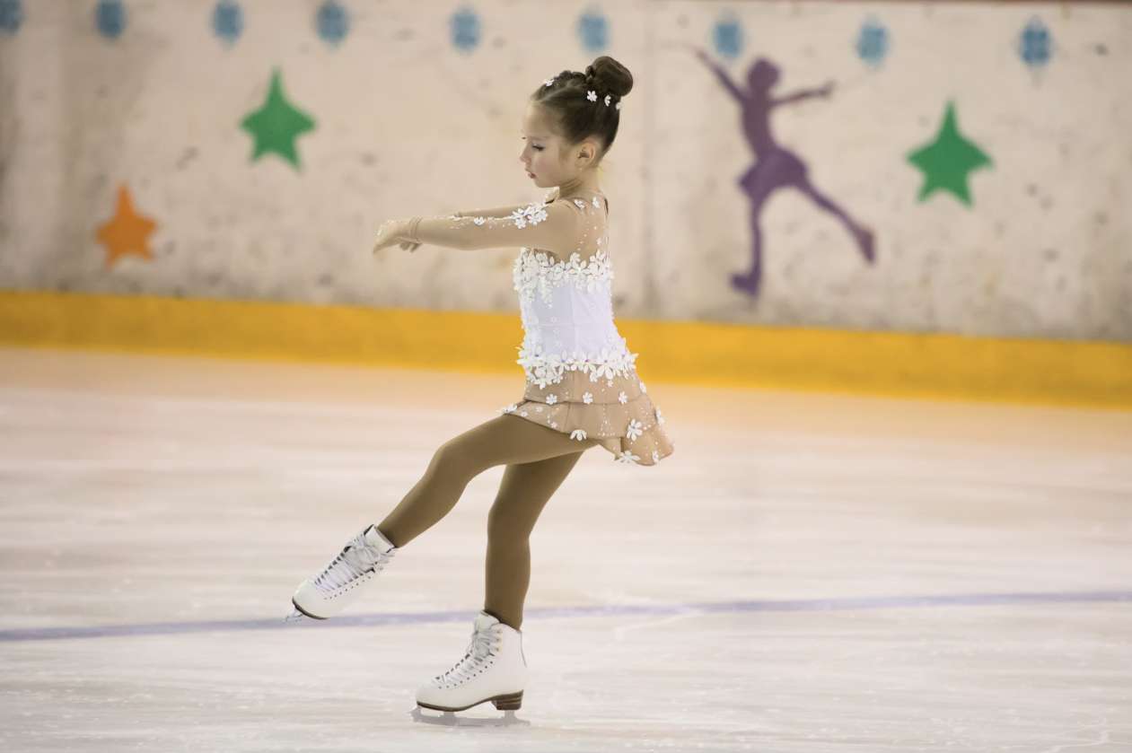 A young female figure skater skates on an indoor ice rink, wearing a white skating costume.