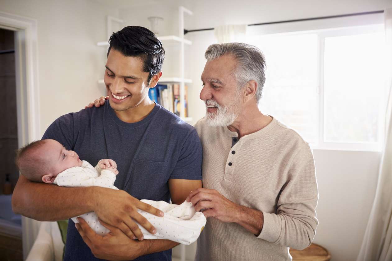 A father holds his newborn baby, while his father stands next to him, with a supportive hand on his arm.