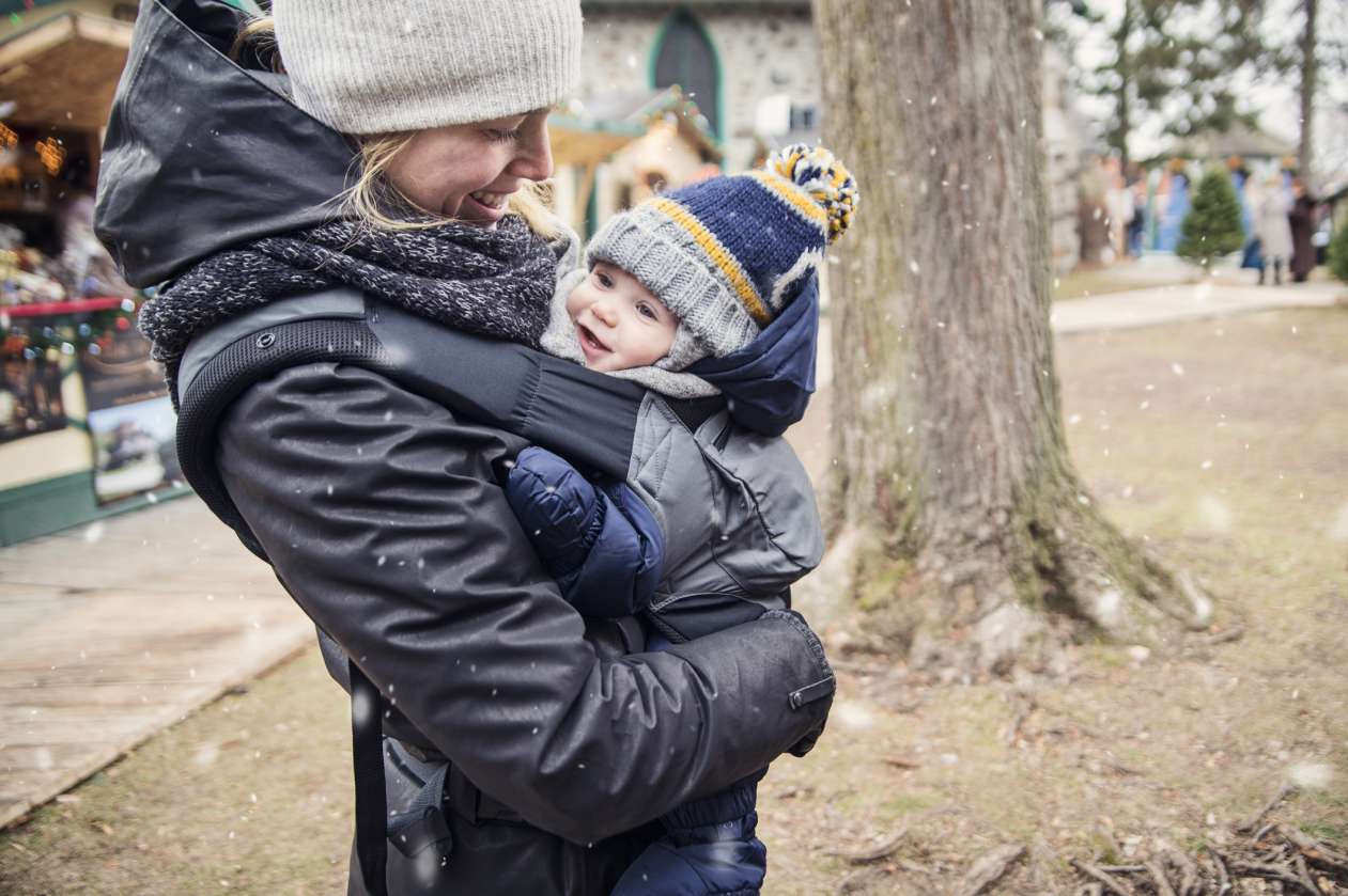 A mom wears her baby in a carrier on her chest. Both are dressed warmly, outdoors in a park as snow falls.