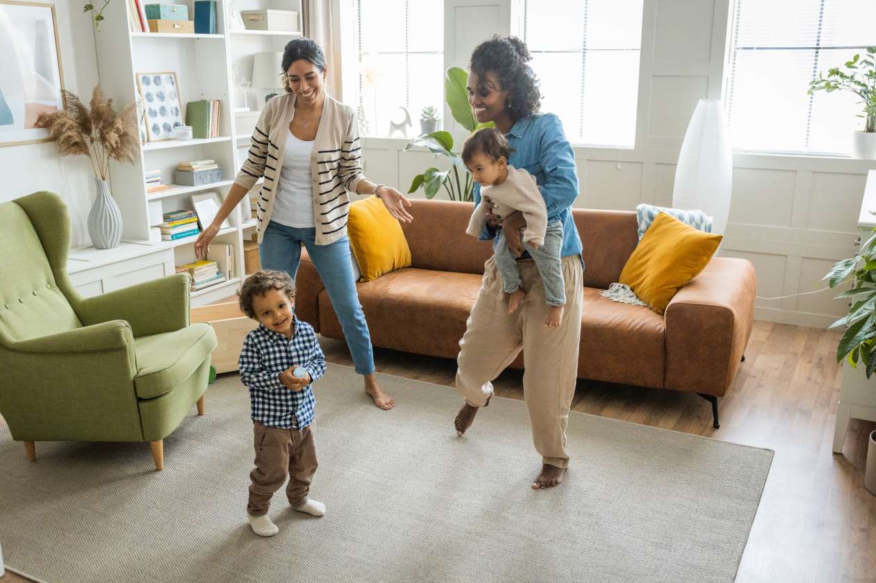 Two mothers dance in the living room with their toddler. One mom holds their baby.