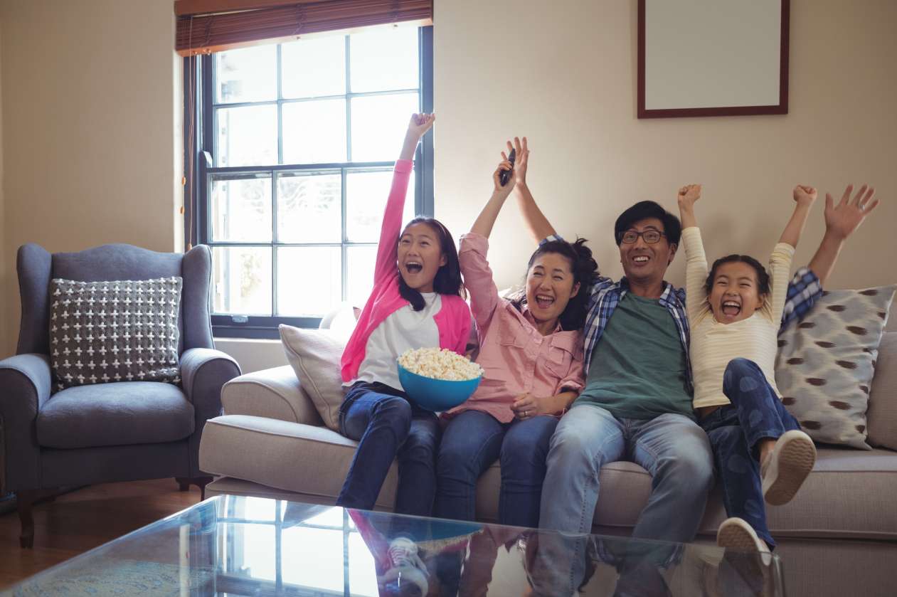 A father, mother, and their two daughters cheer as they watch TV together in their living room.