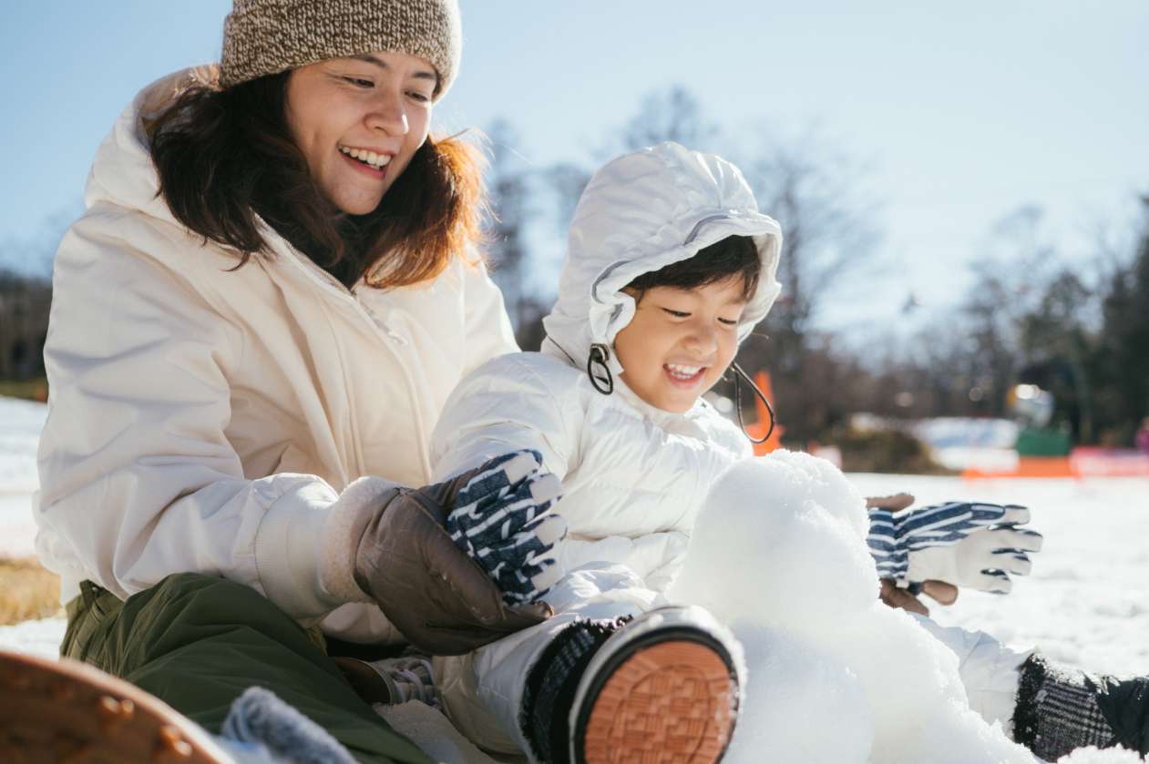 A mother and her young son build a snowman together outside. Both are smiling and sitting on the ground in the sun.