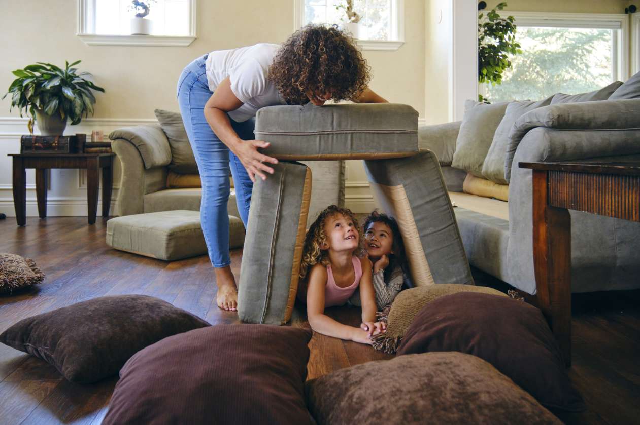 A mother and her two daughters build a fort out of sofa cushions in the living room.