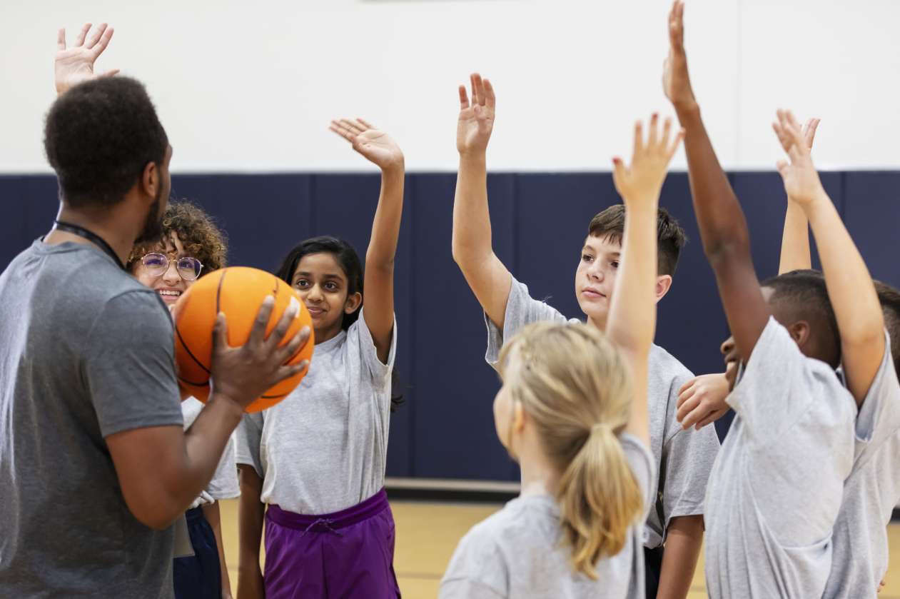 A group of kids in a gymnasium gathers around their PE teacher, who's holding a basketball.