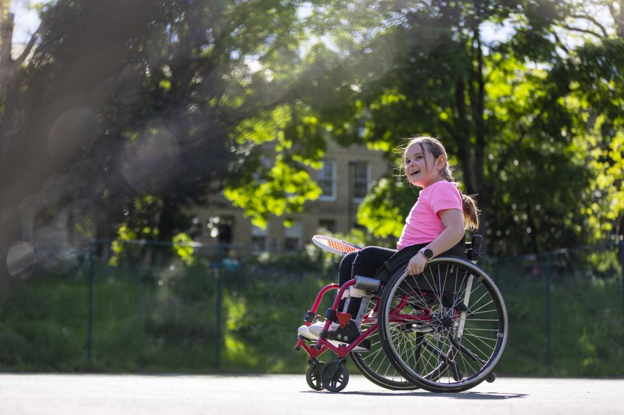 A smiling girl in a wheelchair plays tennis outside on a sunny day.