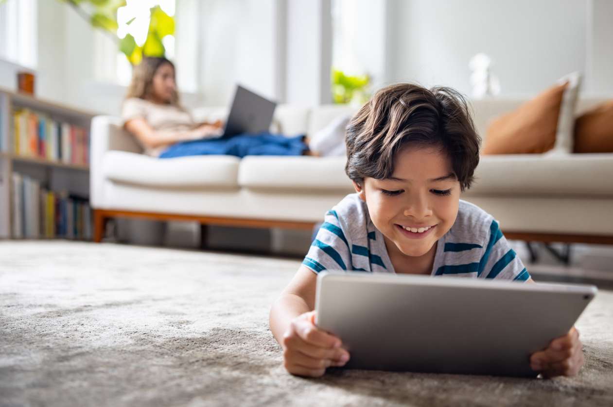 A son lies on his stomach on the living room floor and uses a tablet, while his mom uses a laptop on the couch behind him.