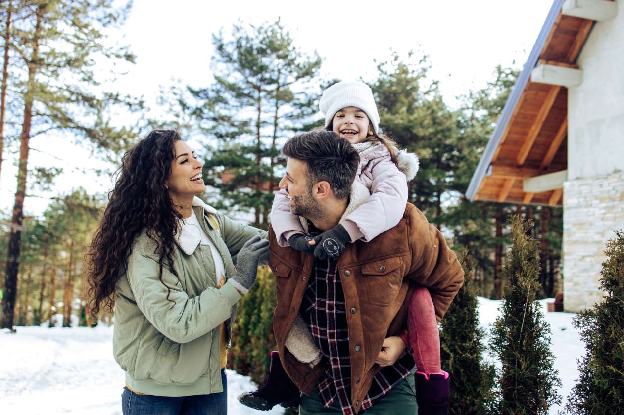 A mother, father, and daughter laugh outside their house together on a winter day. The daughter sits on her dad's shoulders.