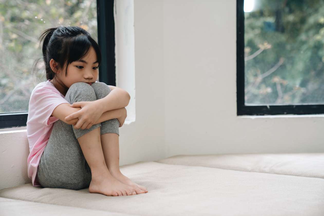 A girl sits on the couch at home, hugging her legs into her chest. She looks sad.