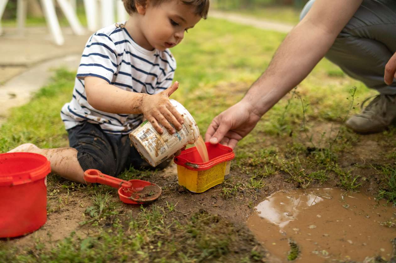 A toddler measures and pours dirt and water outside, helped by his father. Playing with mud and dirt can be a great STEM activity for kids.
