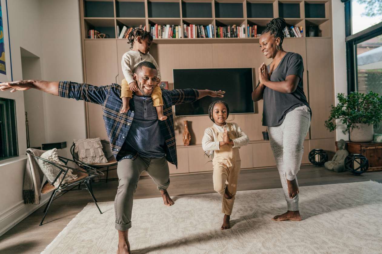 A family does yoga together in the living room. The mother and young daughter do Tree Pose, while the father does a lunge with his toddler sitting on his shoulders.