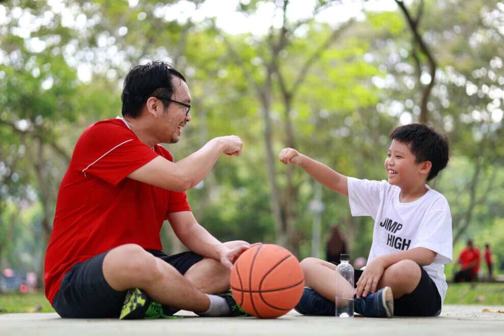 A father and son sit cross-legged outside, with a basketball on the ground between them. They're fist-bumping and smiling at each other.