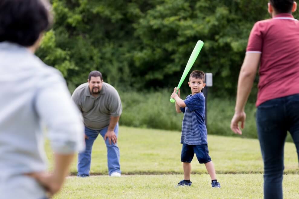 A boy plays scrub baseball in the park with his family.
