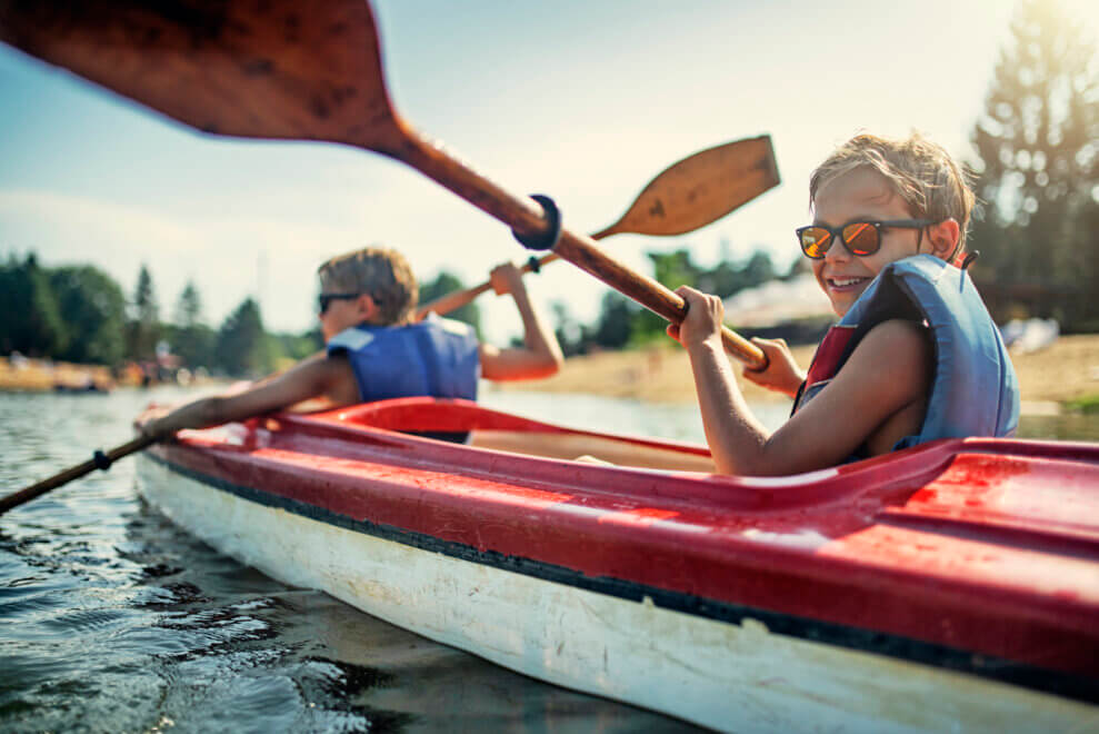 Two brothers paddle together in a kayak on a lake while the sun shines on them and they smile