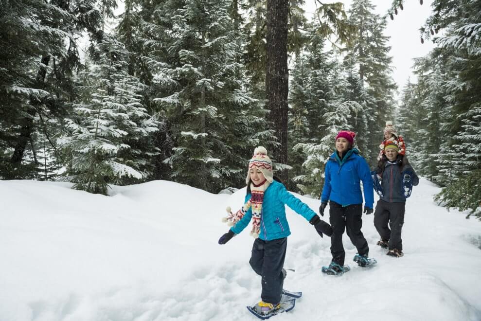 A family with kids walks in the woods while wearing snowshoes.