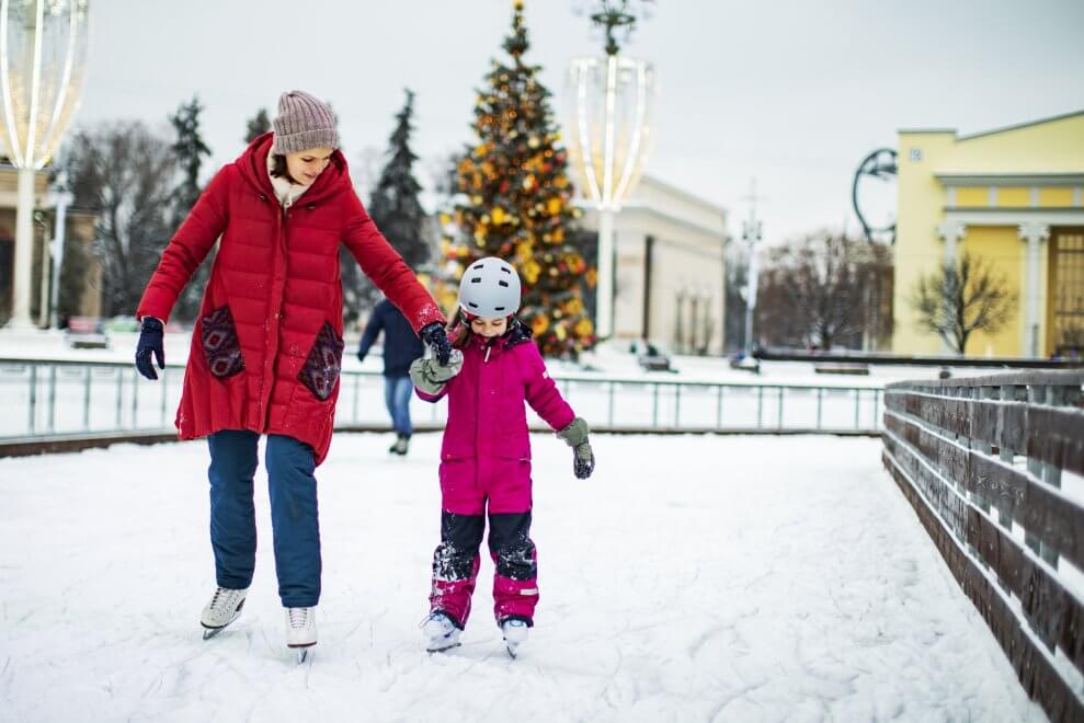 A mother and her daughter ice skate outdoors hand in hand.