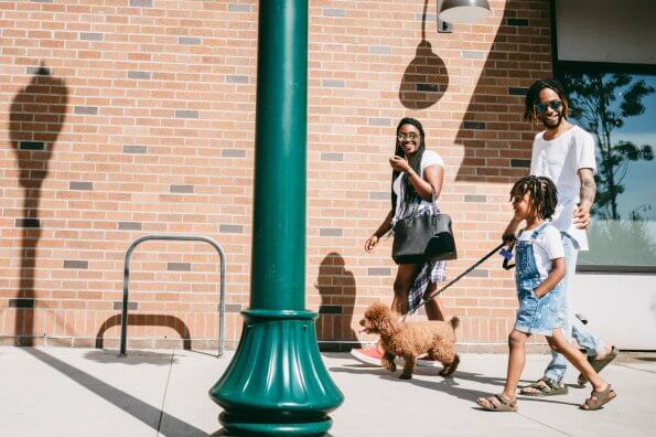 A family consisting of a mother, father, and young daughter walk down a city sidewalk together. The dad walks the family dog on a leash.