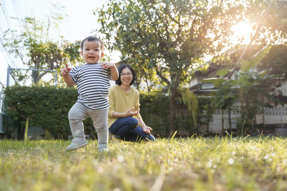 A toddler runs through the grass, as his mother sits behind him, watching. The sun is shining and there are trees behind them.