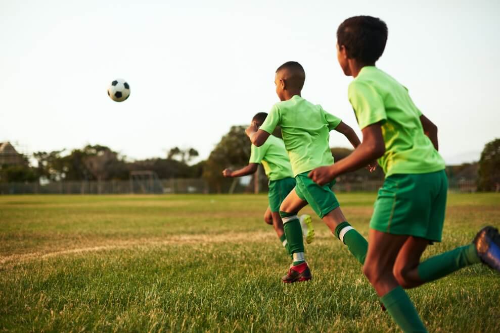 Three boys run after a soccer ball on a sunny evening.