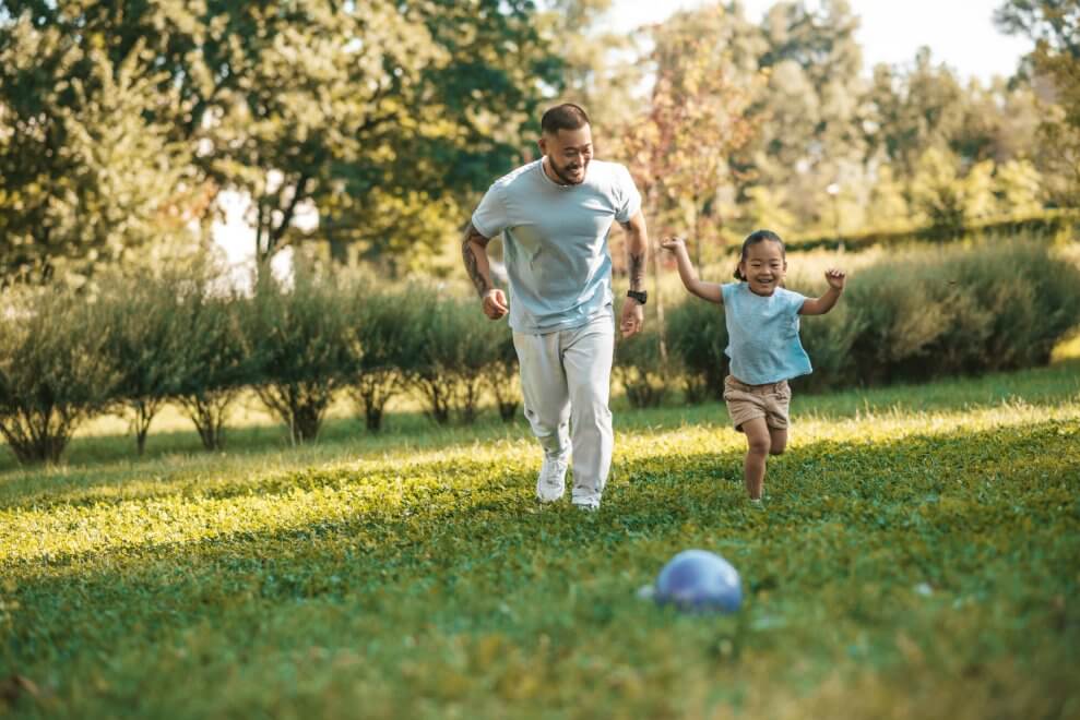 A dad and his daughter run to a ball in a grassy park. They're both laughing. Physical literacy is when kids develop the skills, confidence, and love of movement to be active for life.