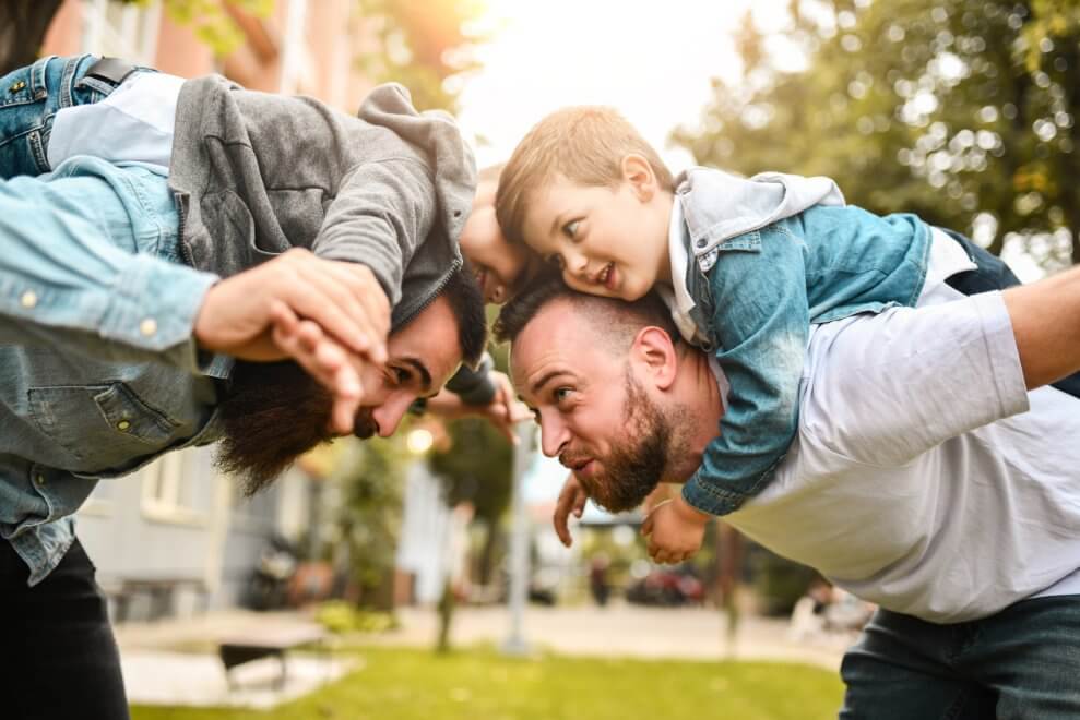 Two dads piggyback and play with their twin sons outside.