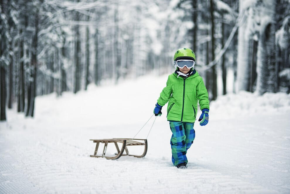 A boy wearing a helmet and goggles pulls his toboggan behind him as he walks in a snowy forest.