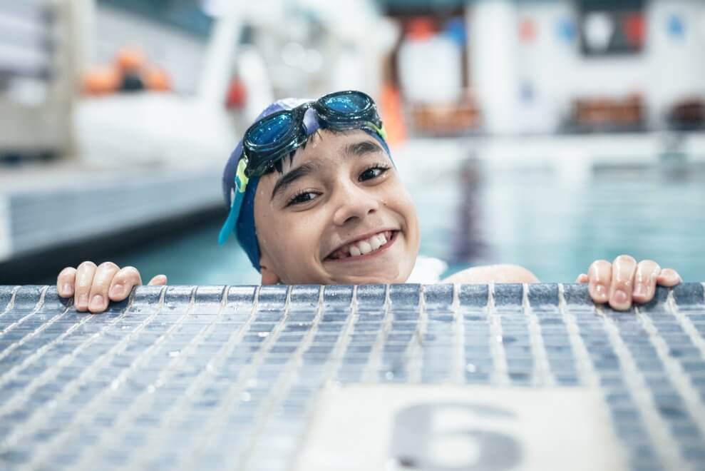 A boy wearing a swim cap and goggles grips the side of the pool, smiling. Swimming is the first event in a triathlon.