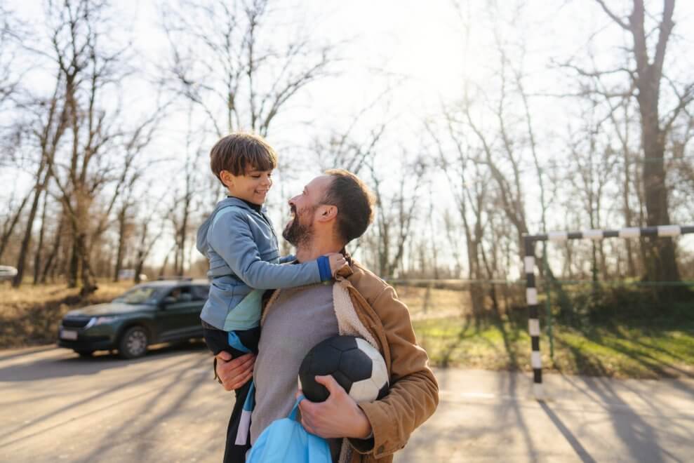 A dad holds his son in his arms, while he clutches a soccer ball. They smile at each other.