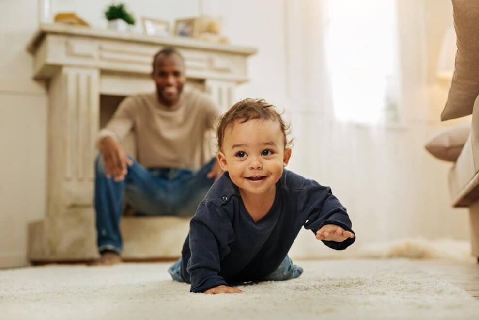 A baby boy crawls across the living-room floor while his dad looks on, smiling.