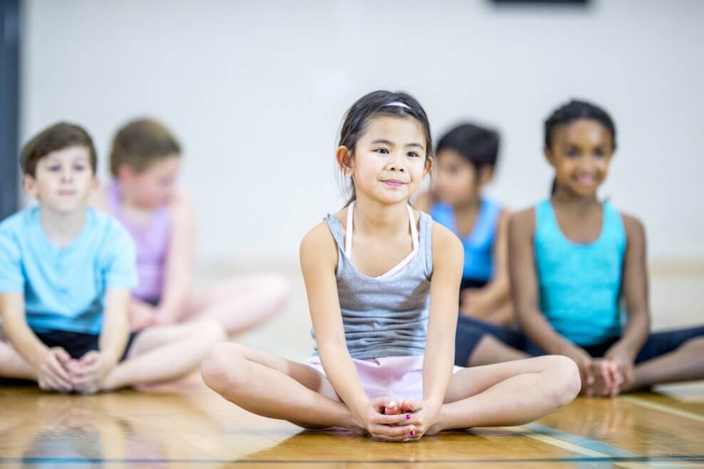 A group of children in a gymnasium sit in butterfly pose.