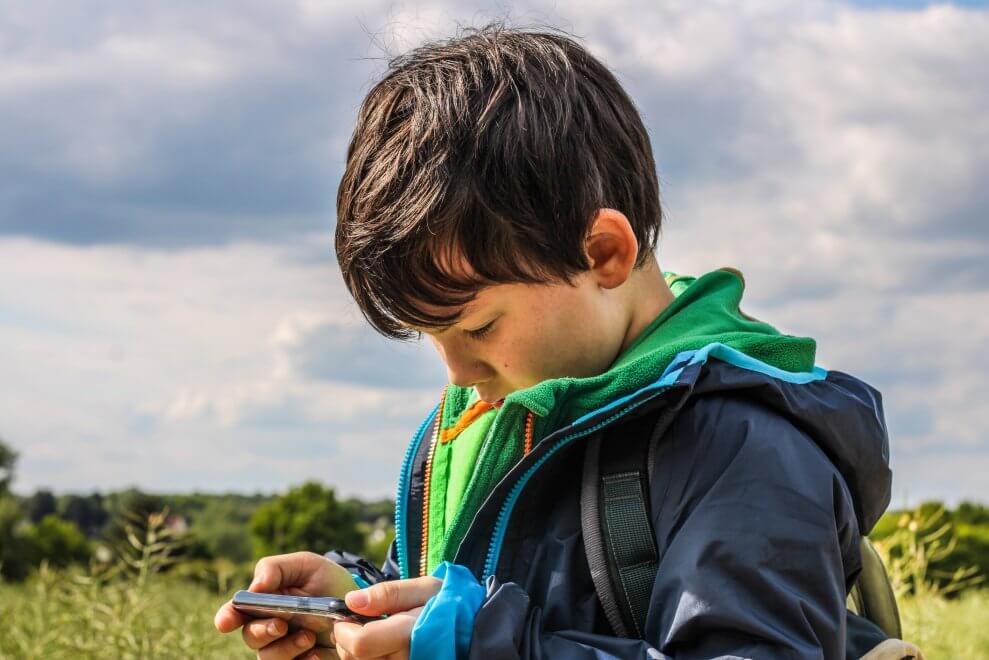 A young boy looks at a cell phone as he geocaches outside on a sunny day.