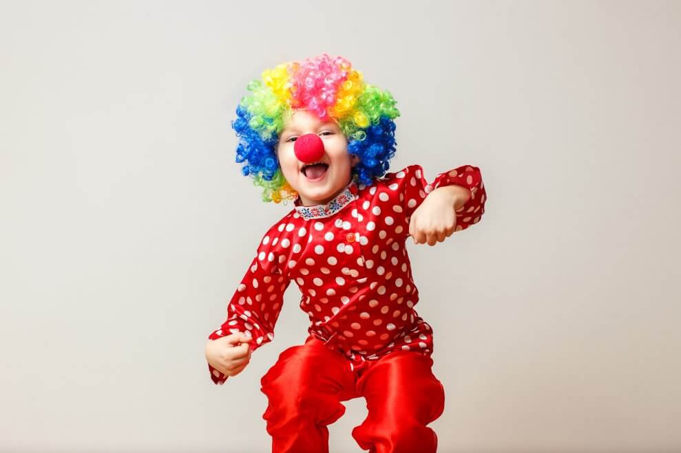 A child is dressed up as a clown, wearing a multi-coloured curly wig, red nose, and red outfit.