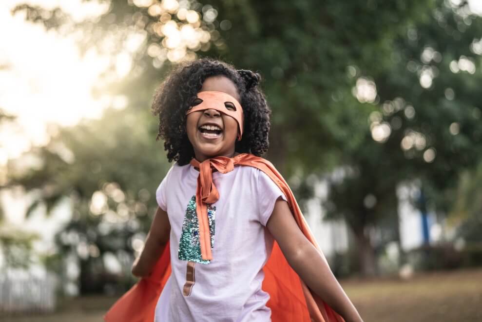 An elementary-age girl dresses up as a superhero. She wears a cape and a mask over her eyes.