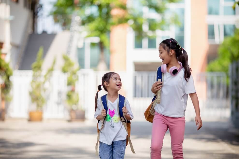 Two sisters walk to school together, laughing, on a sunny day.