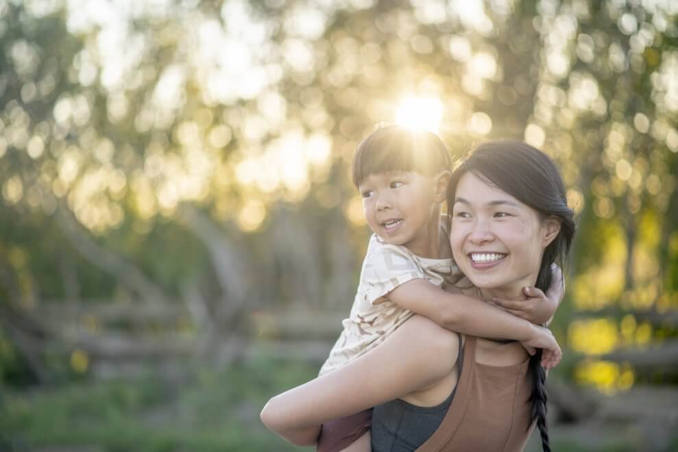 A mother gives her young son a piggy-back ride outside on a sunny evening.