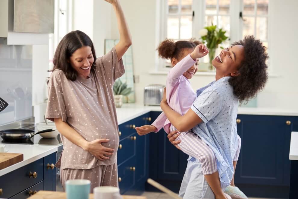 Two moms, one of whom is pregnant, dance in the kitchen in their pajamas. The other mom holds