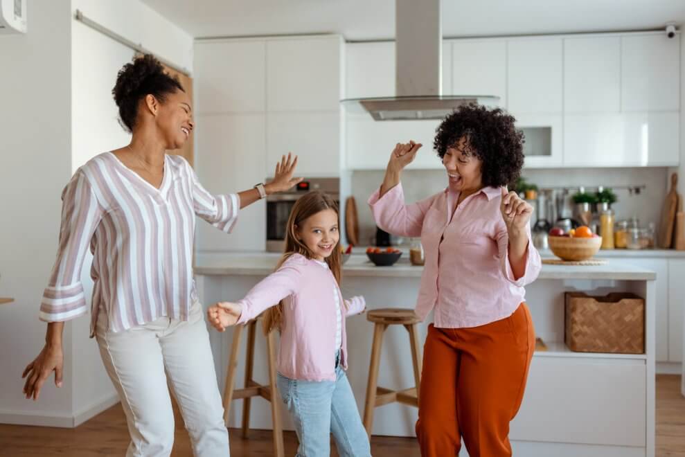 Two moms dance in the kitchen with their elementary-age daughter.