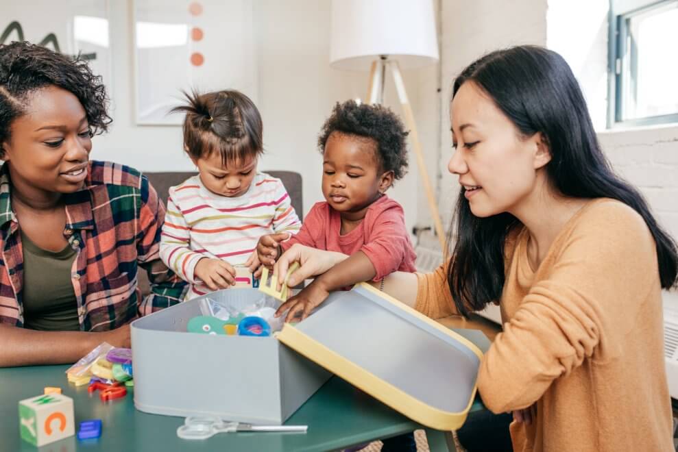 Two mothers play with toys at a coffee table with their toddler daughters.
