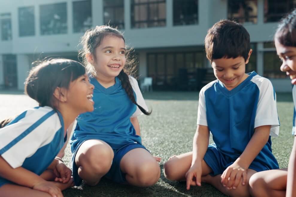 A group of kids wearing soccer jerseys sit on the field and laugh together.