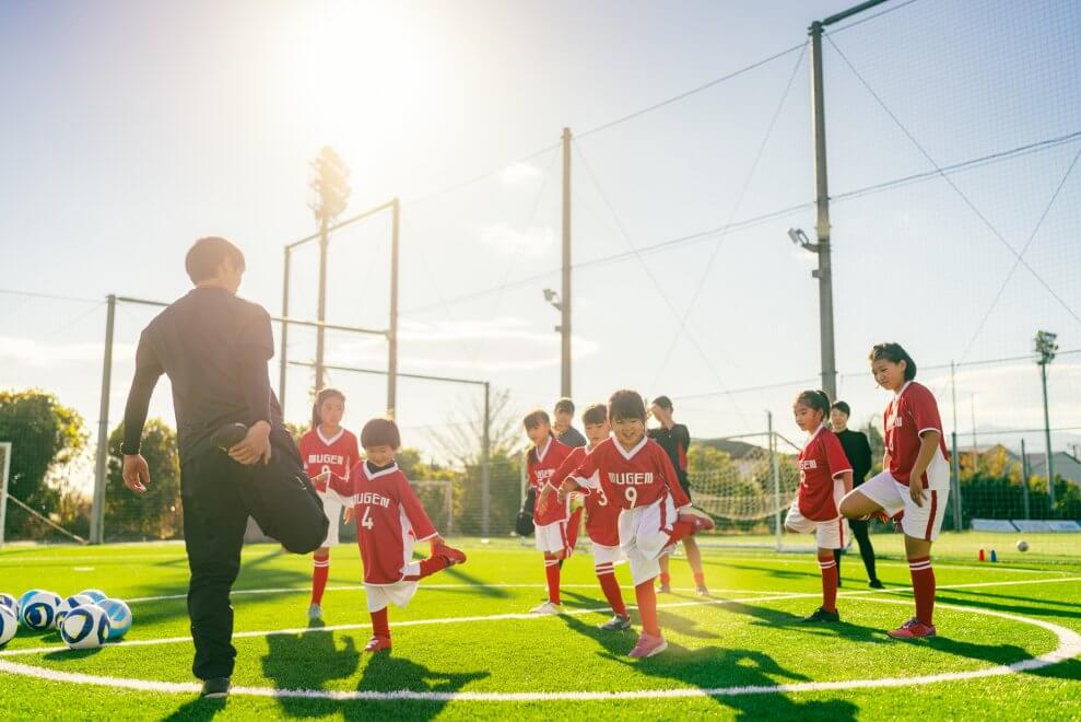 A soccer coach leads his team in a quad stretch on the soccer field on a sunny day.