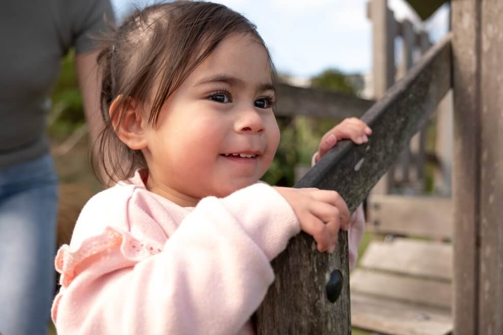 A girl of preschool-age holds onto a railing at a playground outdoors.