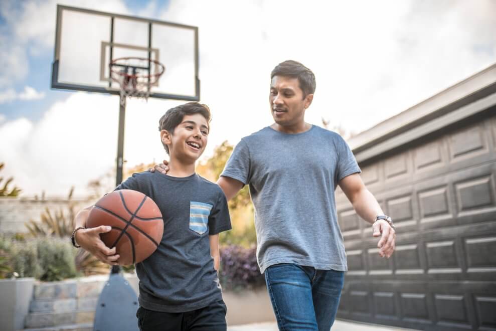 A dad and his son play basketball together in the driveway.