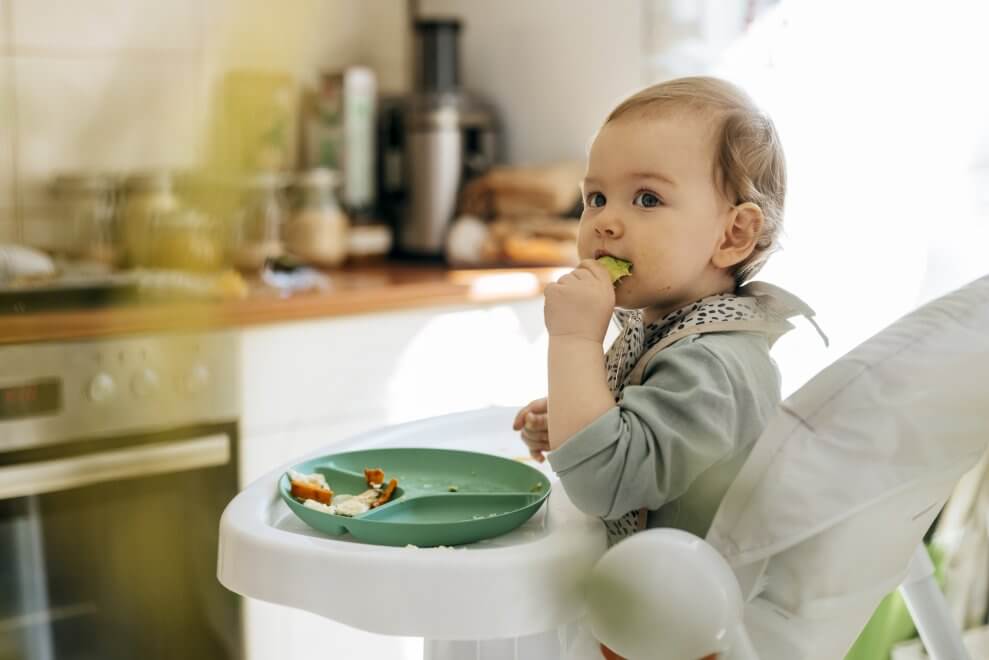 A baby boy sitting in a high chair eats veggies.