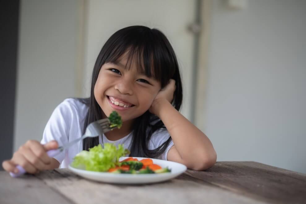A girl sits at the kitchen table eating a plate of veggies with a smile on her face.