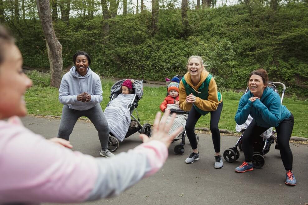 Moms do squats in an outdoor fitness class next to their babies in strollers.