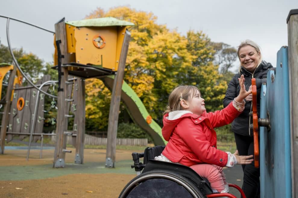 A girl in a wheelchair uses her arms to play with playground equipment as a support person looks on.