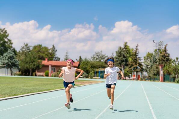 Two smiling girls run on a track underneath a blue sky.