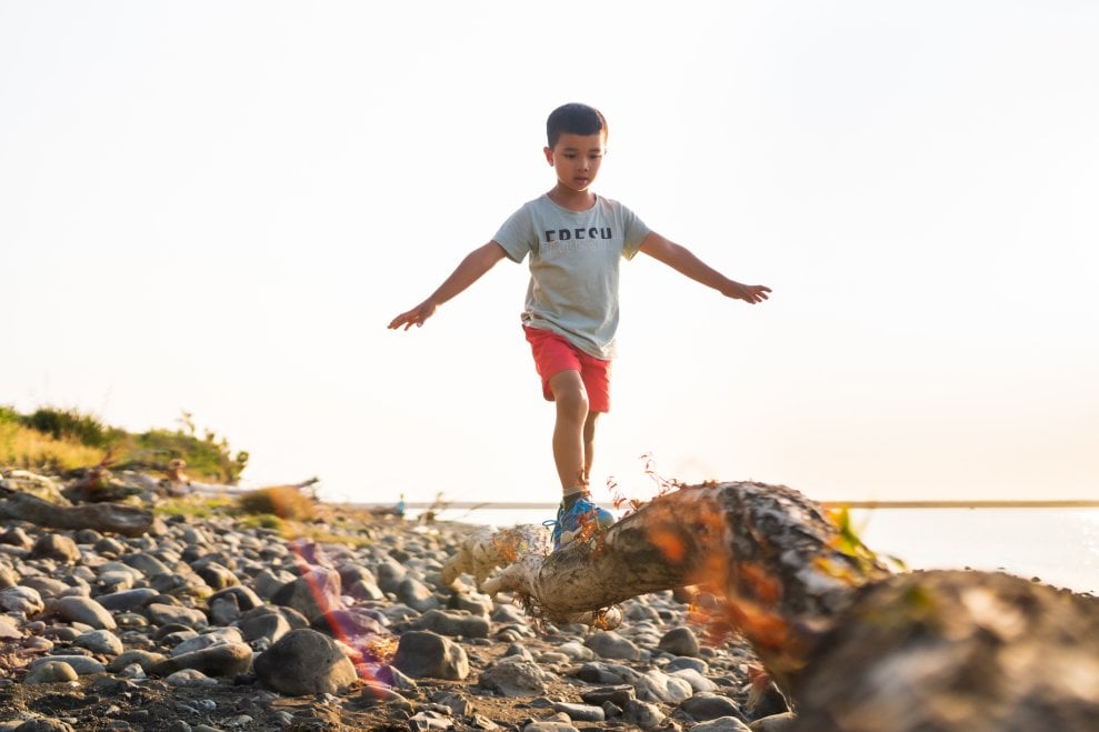 A boy balances as he walks along a log lying on a rocky beach.