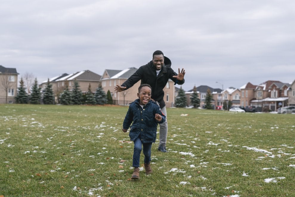 A dad chases his son around a grassy field near a housing subdivision. They're both laughing.  