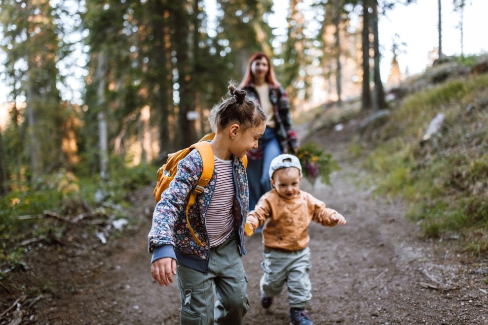 A mother and her two children—an older daughter and a toddler son—hike along a trail in the forest.